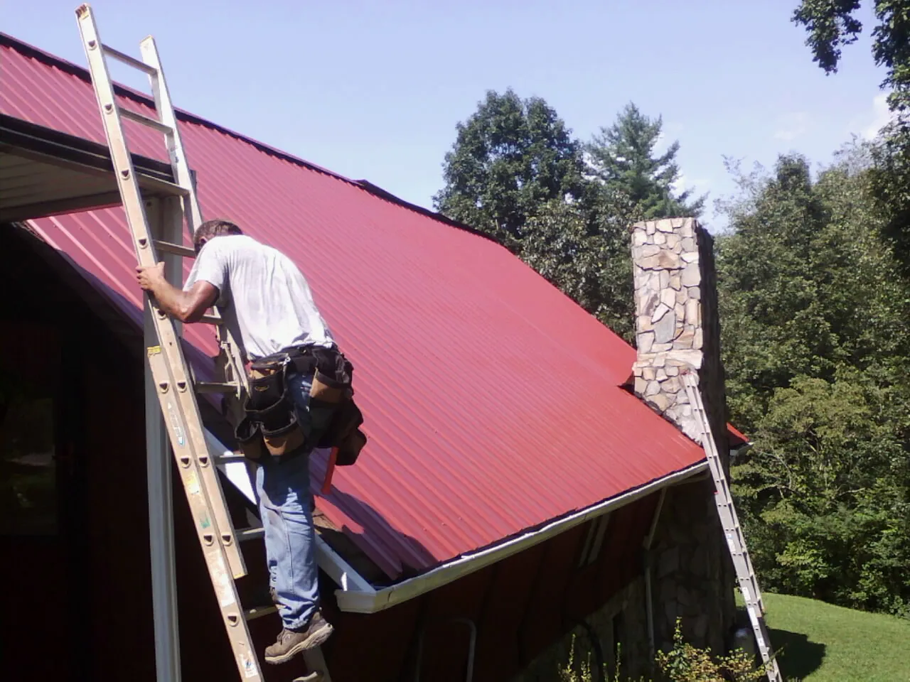 Roofer on a ladder beside a red metal roof and stone chimney — Soffit & Fascia Repair by Roofing Brothers in Morse